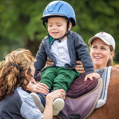 Two volunteers help support a young boy in a blue helmet on the back of a horse